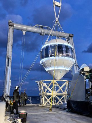 A large white upside-down tear drop shaped space capsule, wrapped at its widest point in tall windows, is suspended over the deck of a sea vessel by a metal grey lift crane. A worker in a helmet stands on the left.