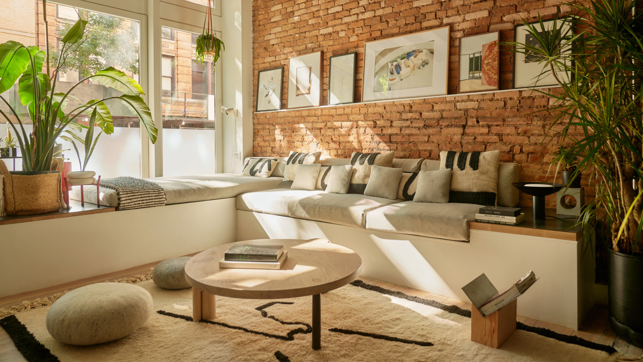 A NYC loft living room with a bench full of neutral cushions, a brick wall with frames, a potted plant, a cream rug, a wood coffee table, and a book stand