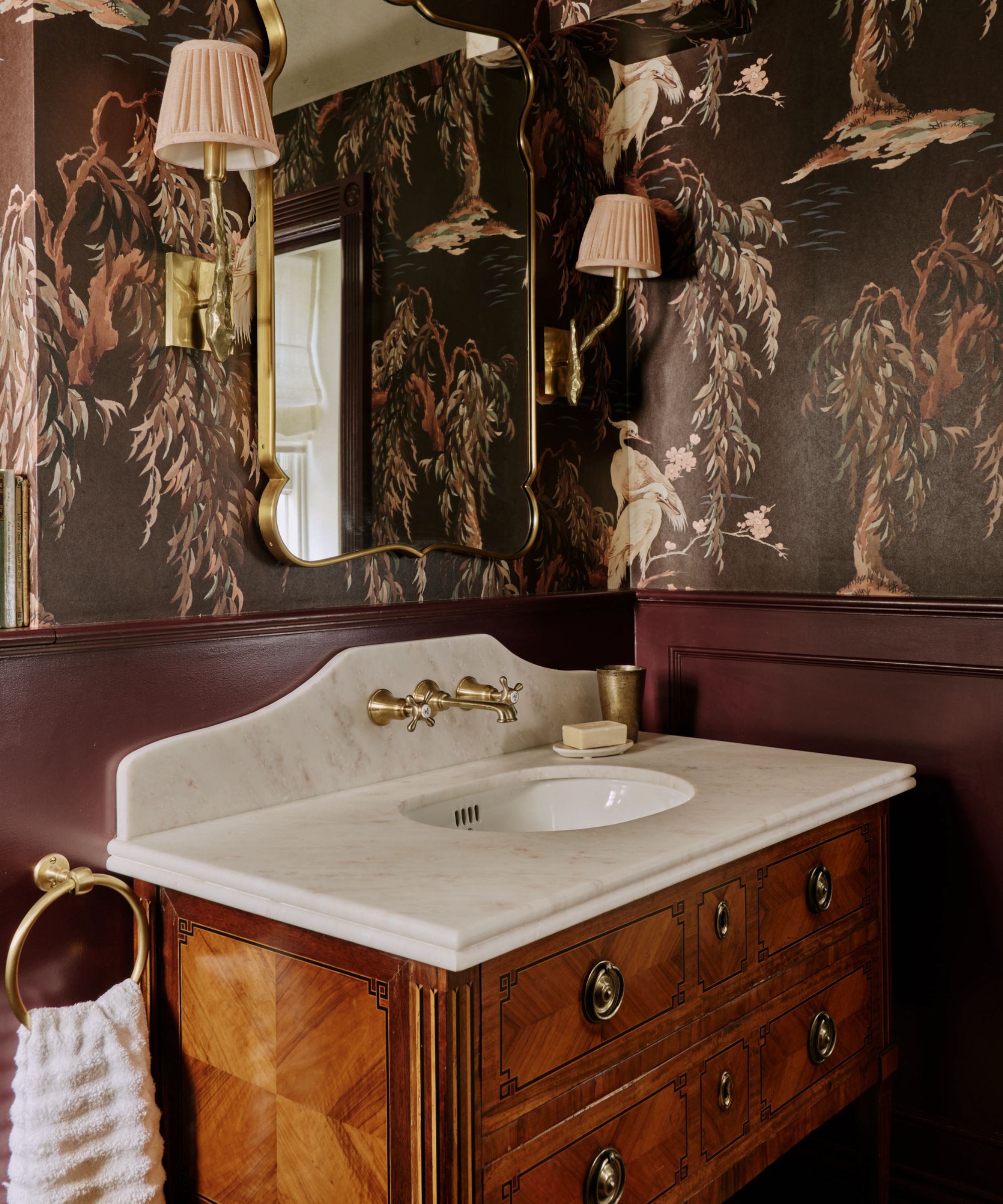 a small dark moody powder room with a chocolate brown wallpaper and half height paneling painted burgundy brown with a antique wood vanity and marble sink