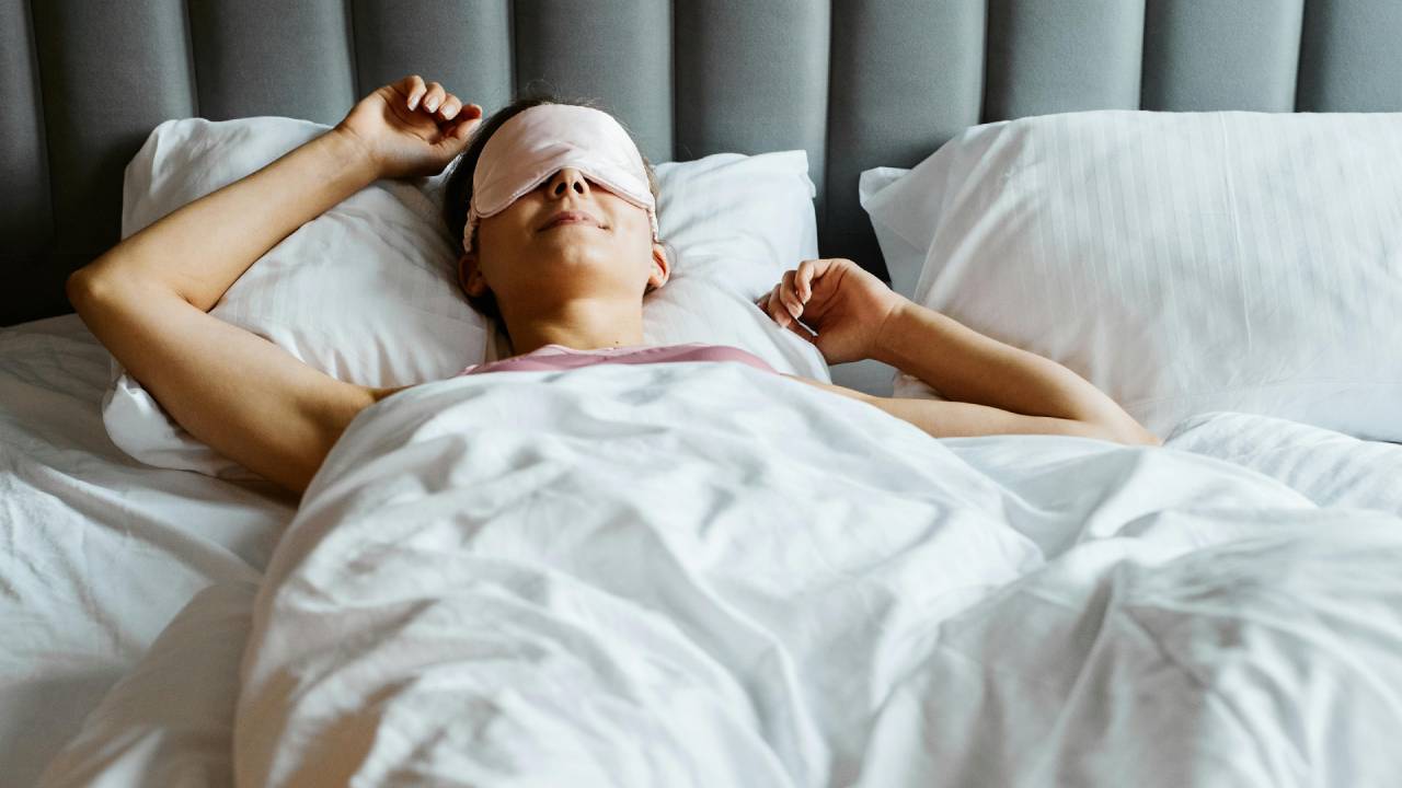 A woman lying on a mattress under white sheets while wearing a light pink eye mask