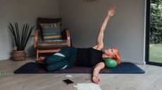 Woman exercising, lying on her side on a mat with a Pilates ball under her head and her right arm raised vertically