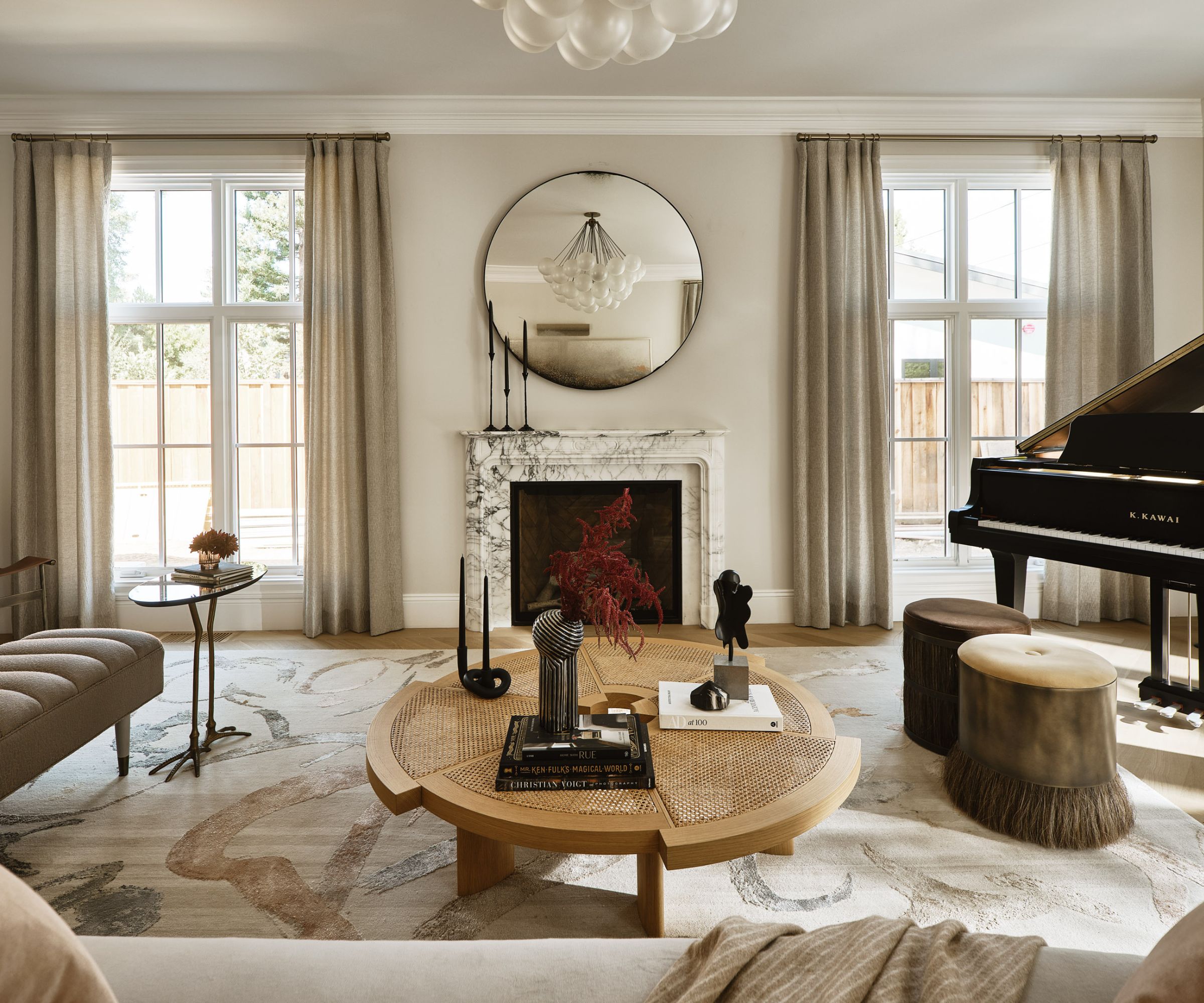 Living room with beige walls, large windows and linen drapes, rattan coffee table, velvet brown stools, cream sofa, large cream abstract rug and piano