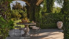 european style garden with stone arches, lush greenery, and a round concrete pedestal table with 4 wrought iron chairs and a bowl of lemons