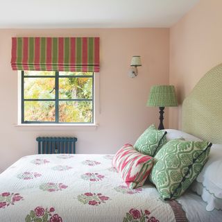 cottage bedroom with pale pink painted walls and bed with green headboard, green lamp, striped red and green roman blind, floral quilt