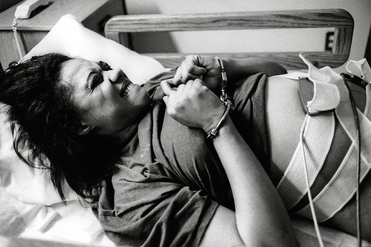 Black and white photograph of a woman handcuffed in a bed with tubes and straps taped to her stomach