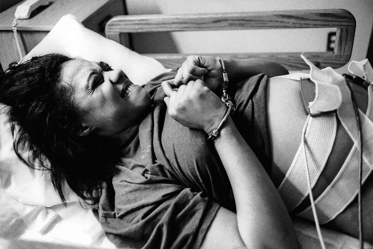 Black and white photograph of a woman handcuffed in a bed with tubes and straps taped to her stomach