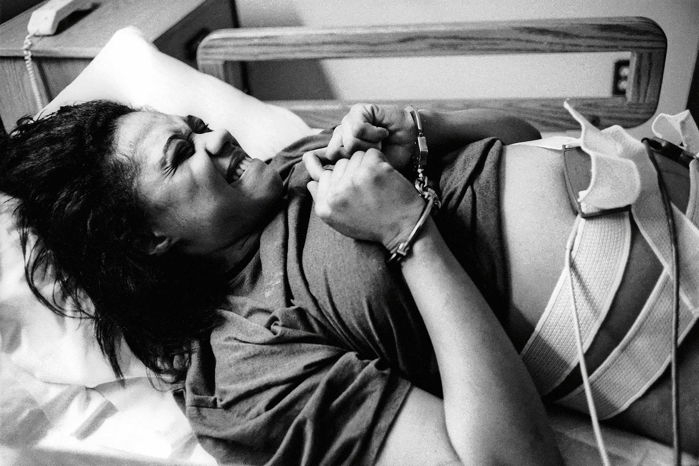 Black and white photograph of a woman handcuffed in a bed with tubes and straps taped to her stomach