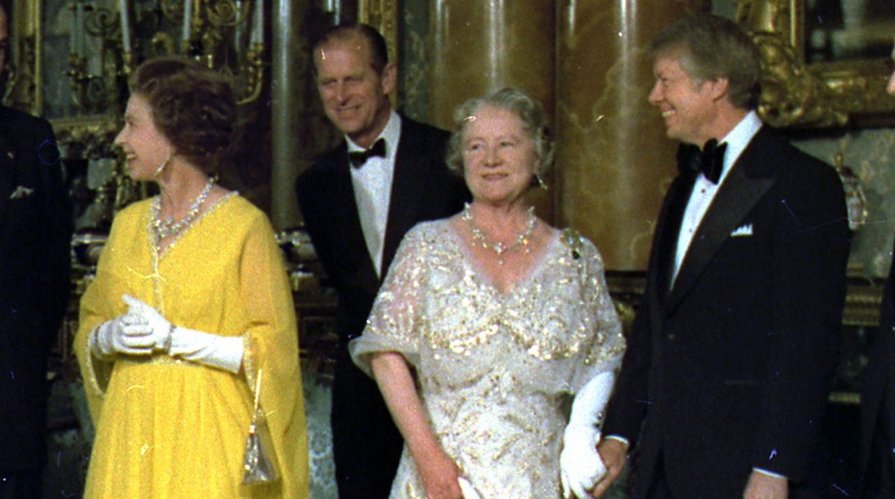 Jimmy Carter wears a tuxedo and holds hands with the Queen Mother, who is wearing a sparkly white and gold gown, alongside Prince Philip, who wears a tuxedo, and Queen Elizabeth II, who wears a bright yellow gown