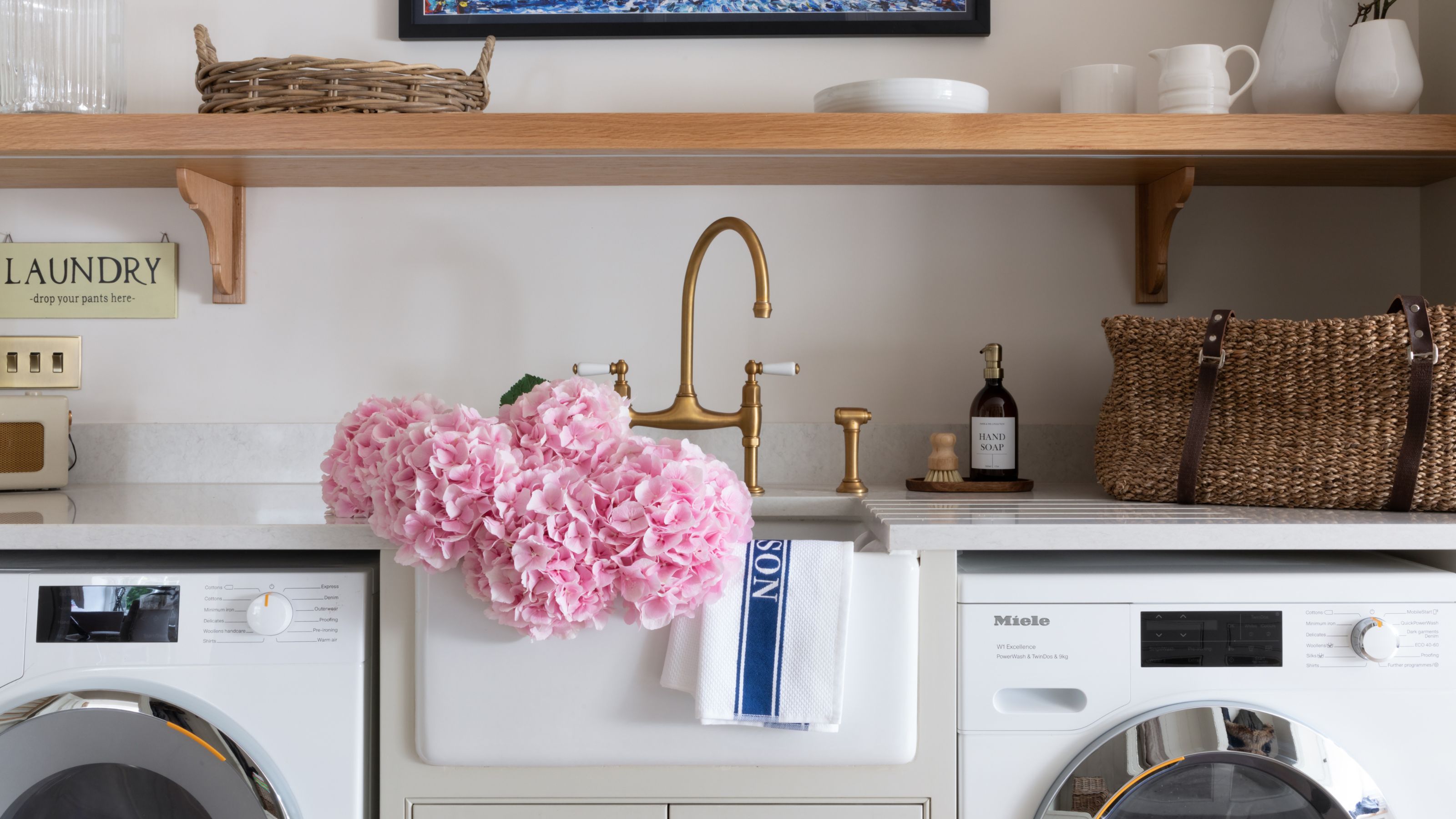 Utility room with a washing machine and tumble dryer next to a white ceramic sink with pink hydrangeas sitting in it