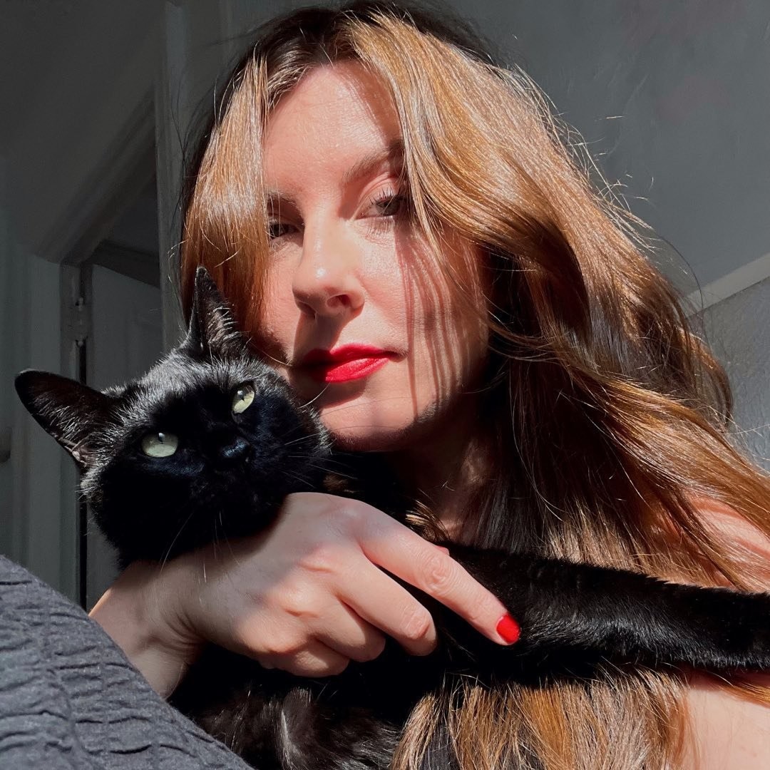 A woman with red lipstick and red nails poses with a black cat.