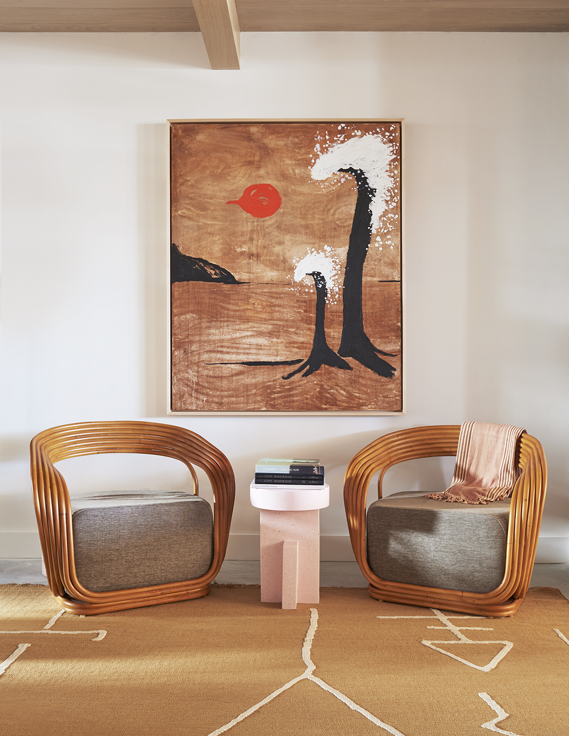A hotel lounge is decorated with sculptural, abstract furniture and artworks in earthy, terracotta hues, including a rug, two matching rattan and textile armchairs, a pink stool-turned-coffee-table, and a large painting.