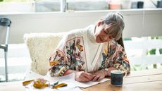 An older woman smiles as she writes in a notepad at her kitchen table.