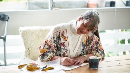 An older woman smiles as she writes in a notepad at her kitchen table.
