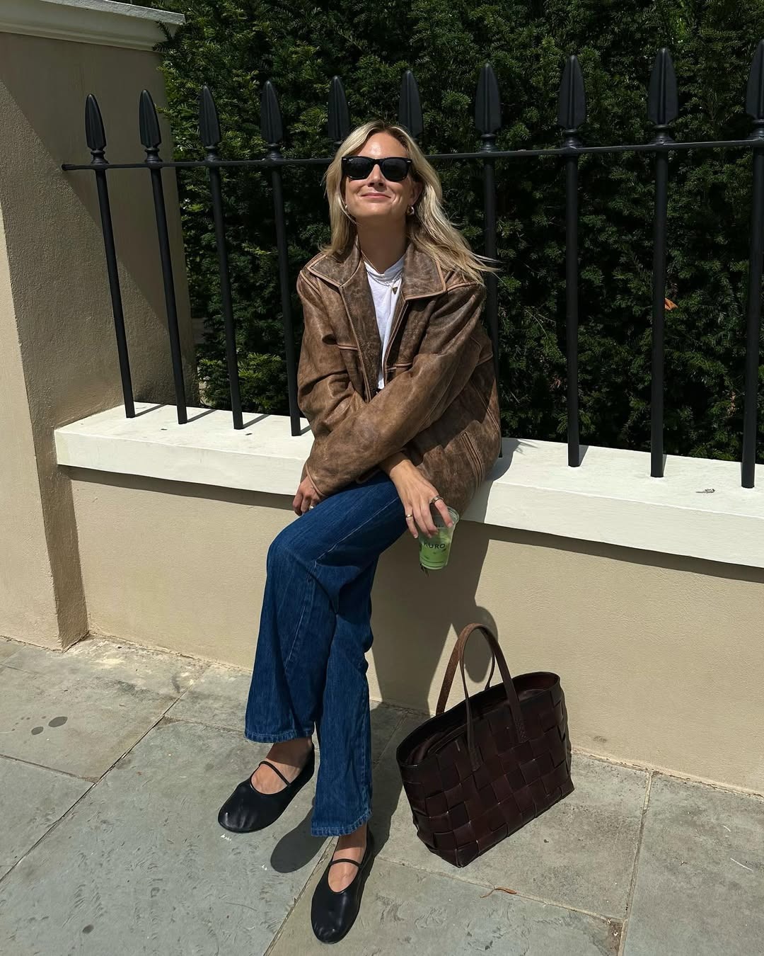 Woman wears brown leather jacket, white t-shirt, blue jeans, ballet flats, brown knitted leather bag