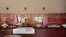 A deep burgundy colored kichen with a Belfast sink, shown with a mix of plain and subtle textured tiles forming the backsplash. 