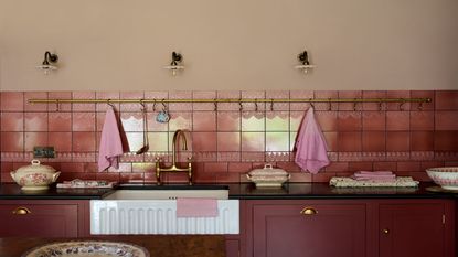 A deep burgundy colored kichen with a Belfast sink, shown with a mix of plain and subtle textured tiles forming the backsplash. 