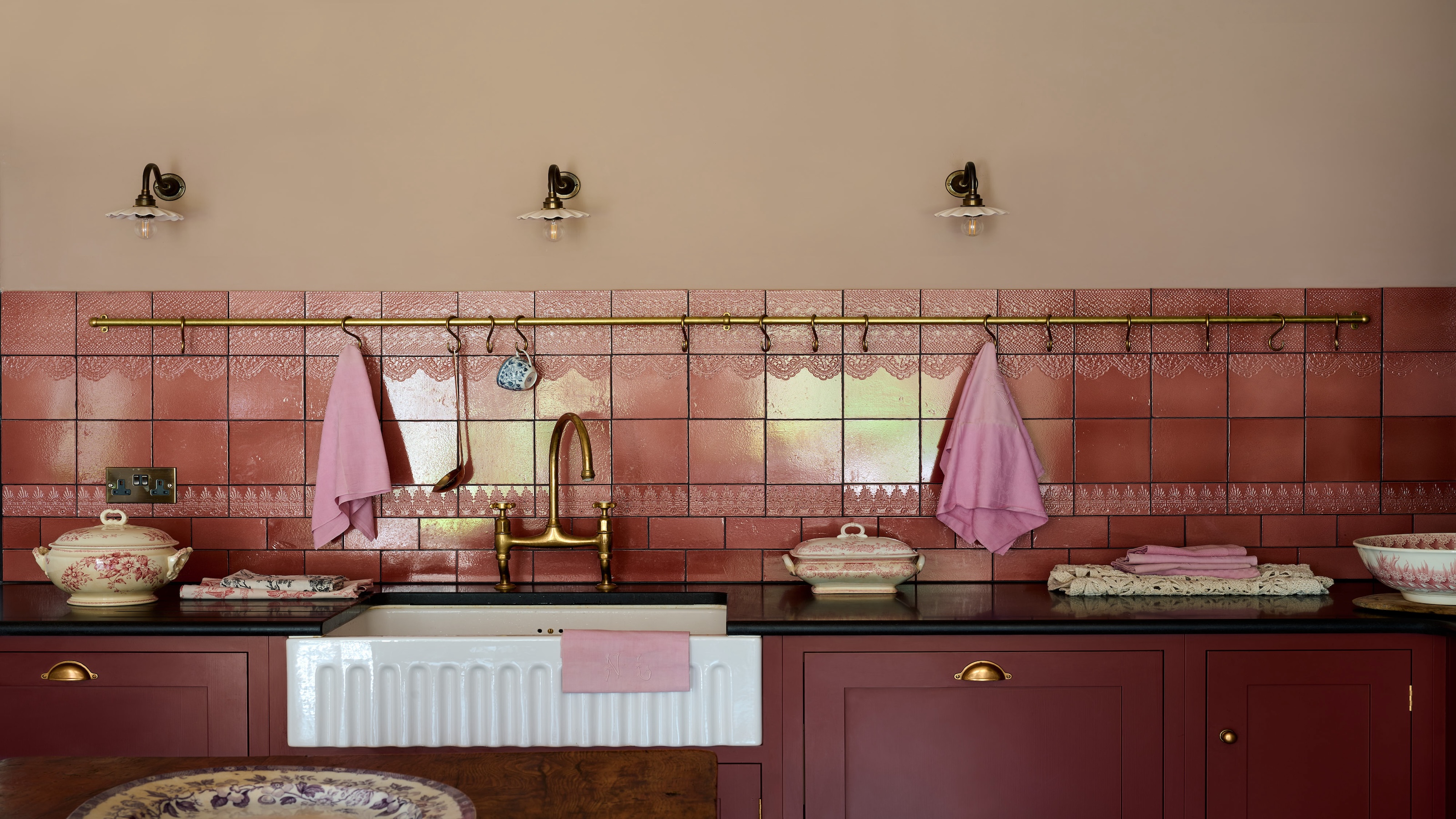 A deep burgundy colored kichen with a Belfast sink, shown with a mix of plain and subtle textured tiles forming the backsplash. 