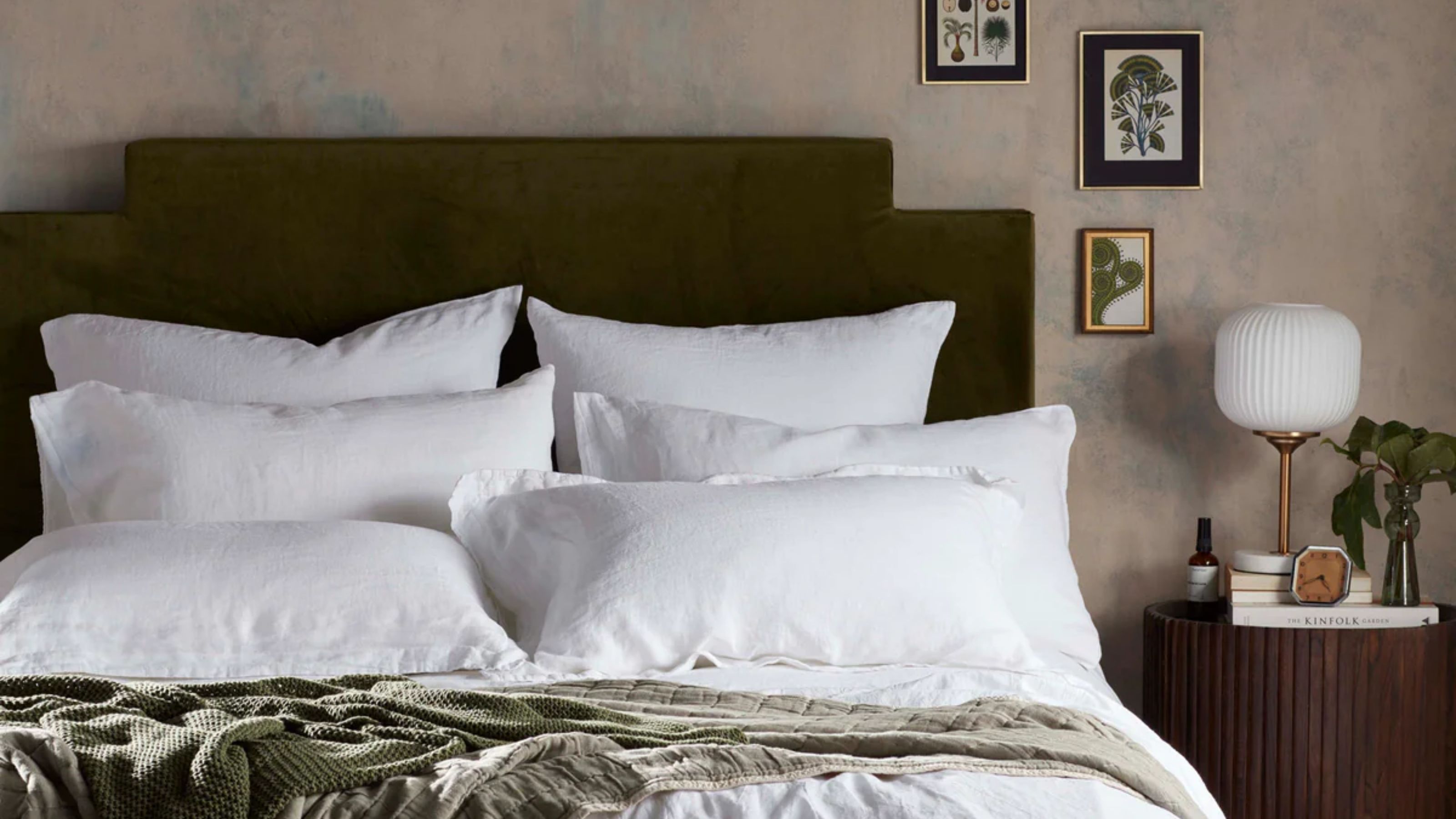 Image of a bed with a green velvet headboard and white bedding. There are six white pillows on the bed and a sage green throw blanket at the end of the bed.