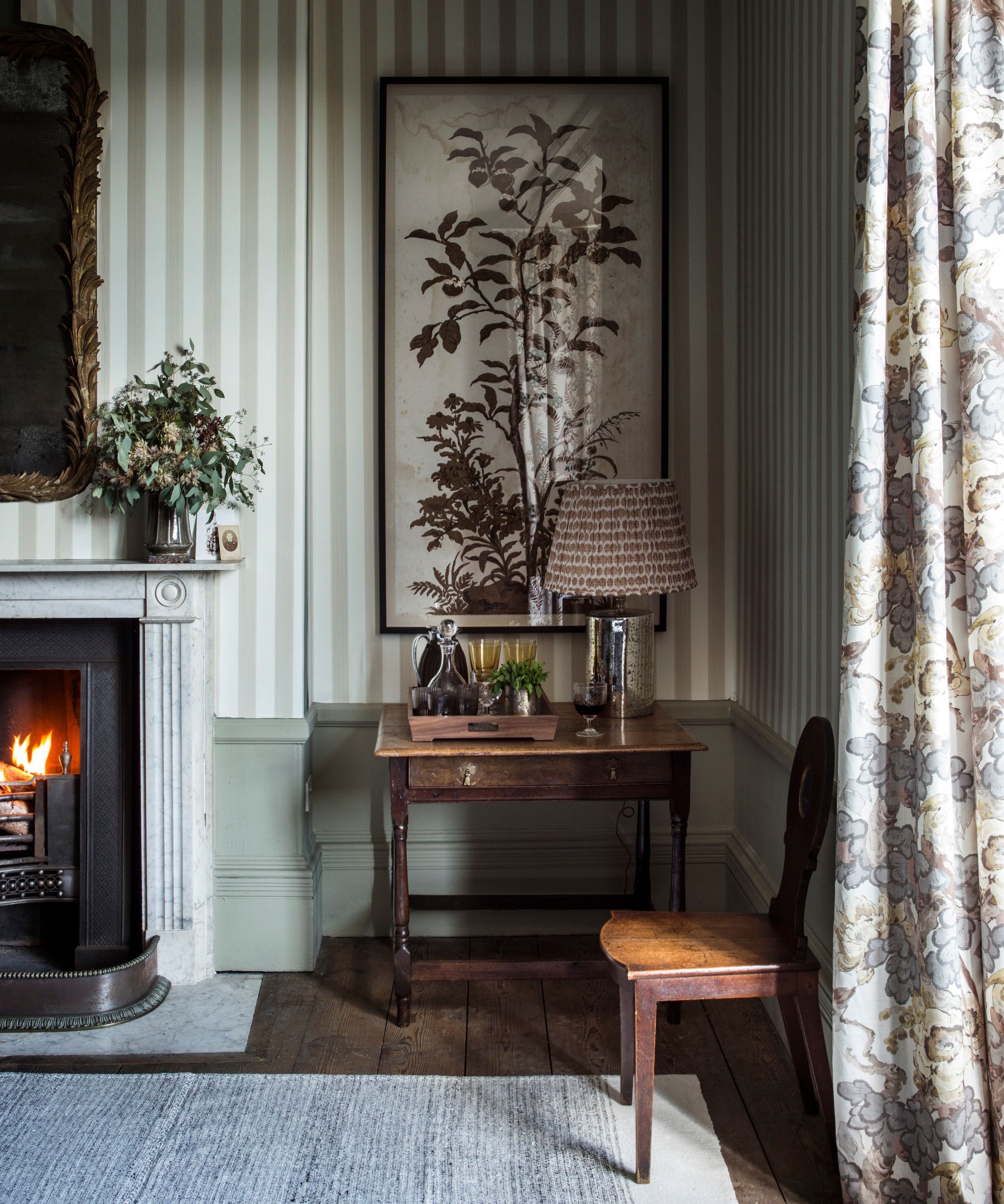 A cozy autumnal living room with striped wallpaper, a lit fire, artwork, floral curtains and a large gold-framed mirror. The flooring is wooden with a grey rug on top.
