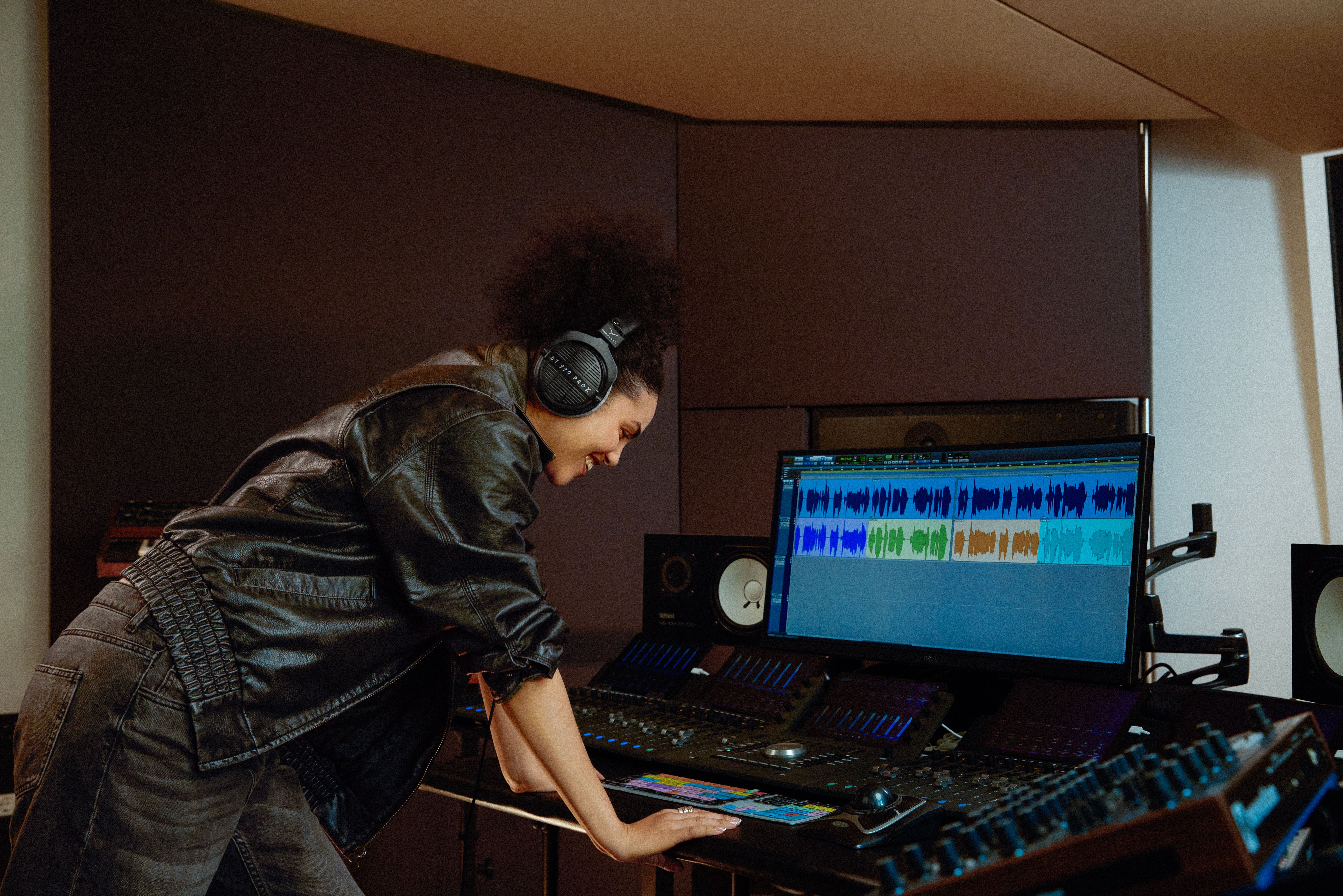 Lifestyle image for the Beyerdynamic DT-990 Pro X headphones. A woman in a black leather jacket leans over a mixing desk, looking at a music app and grinning
