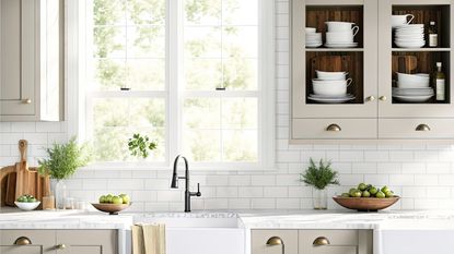 A window over a kitchen farmhouse sink with a black tap, flanked by beige glass front cabinets filled with dinneware