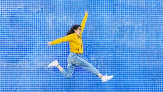 Why is sleep important: A happy woman wearing a yellow jumper and blue jeans jumps in the air