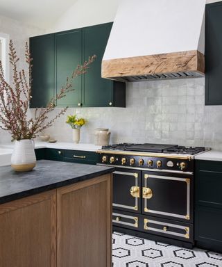 A kitchen with a twin oven Le Cornue range in black and brass, with green cabinets, a reclaimed oak hood and white kitchen backsplash ideas in zellige tiles.