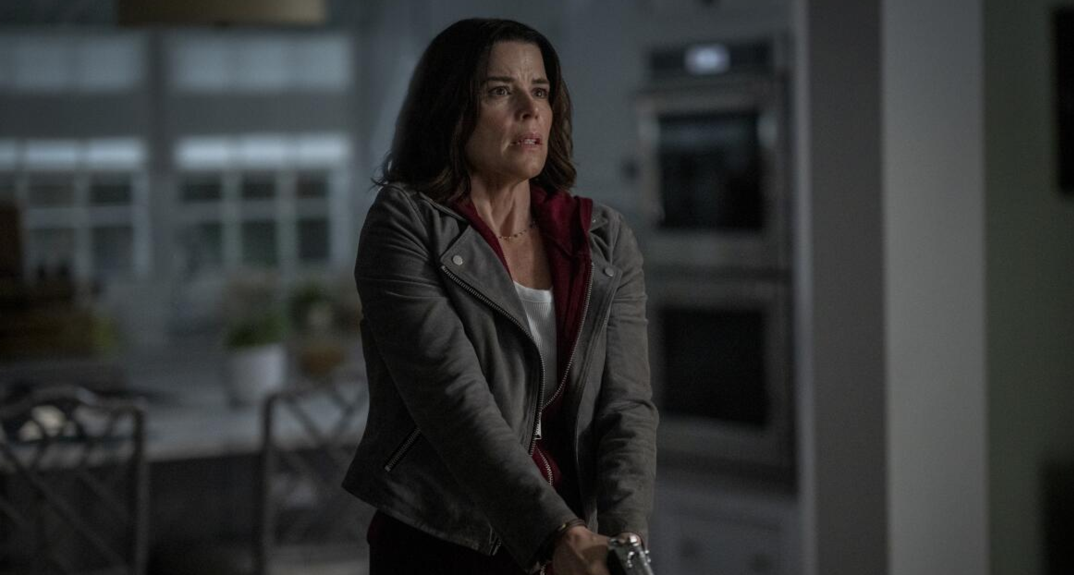 neve campbell as adult sidney prescott looking scared and holding a gun while walking through a dark house in a still from scream 7