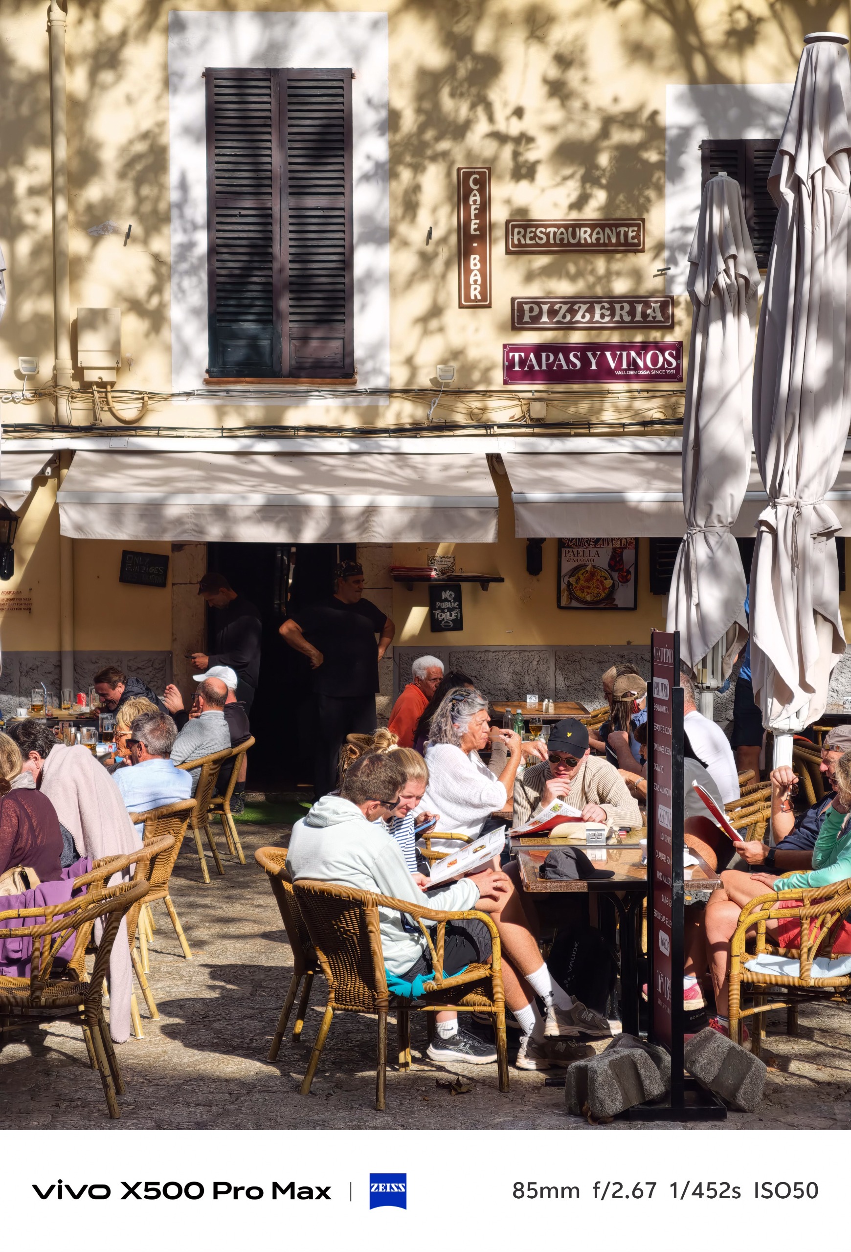 Busy outdoor restaurant terrace beneath signs advertising a cafe, pizzeria, tapas and wine.