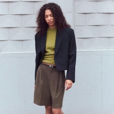 A Zara model with dark, curly hair wears a cropped, boxy blazer, green knit, tailored long brown shorts, and a skinny black belt.