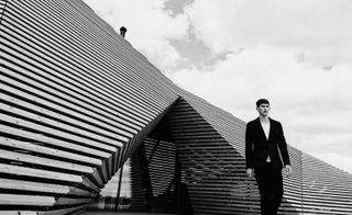 A black and white photo of a man wearing a suit standing in front of a building made of striped panels with a triangular shaped entrance.