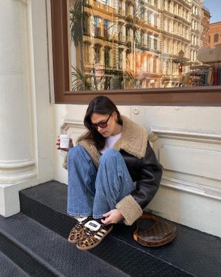 Influencer @maggie_mccormack  wears a pair of leopard print trainers with blue jeans and a black leather jacket whilst drinking coffee on steps