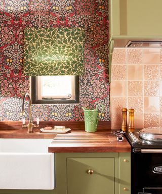 Vibrant kitchen with pink floral patterned wallpaper, green painted kitchen cabinets, green patterned blind on small window, wooden countertop, black aga, salmon pink tiles on wall above.