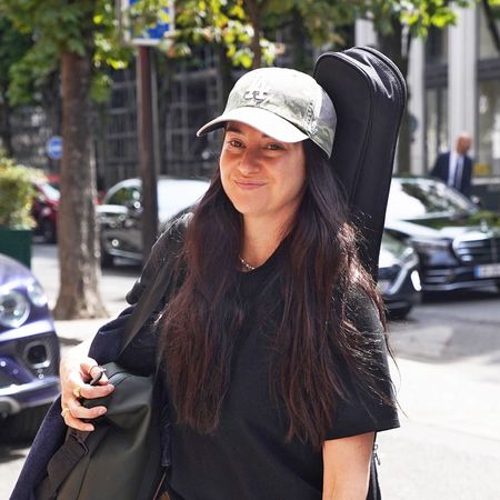 Shailene Woodley wearing a green baseball cap, black t-shirt, olive pants, and black loafers 