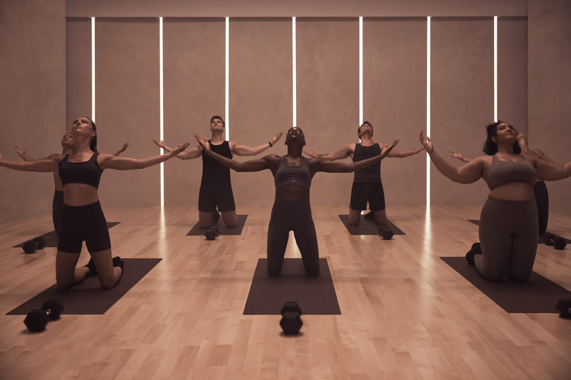 A series of people dressed in black fitness clothing is photographed in a wood-clad pilates studio lit by vertical beams of light.