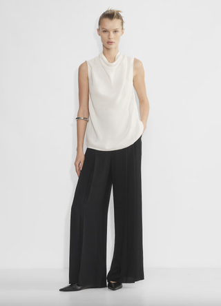 a model in front of a plain backdrop wearing flowy pants and a draped top by aritzia