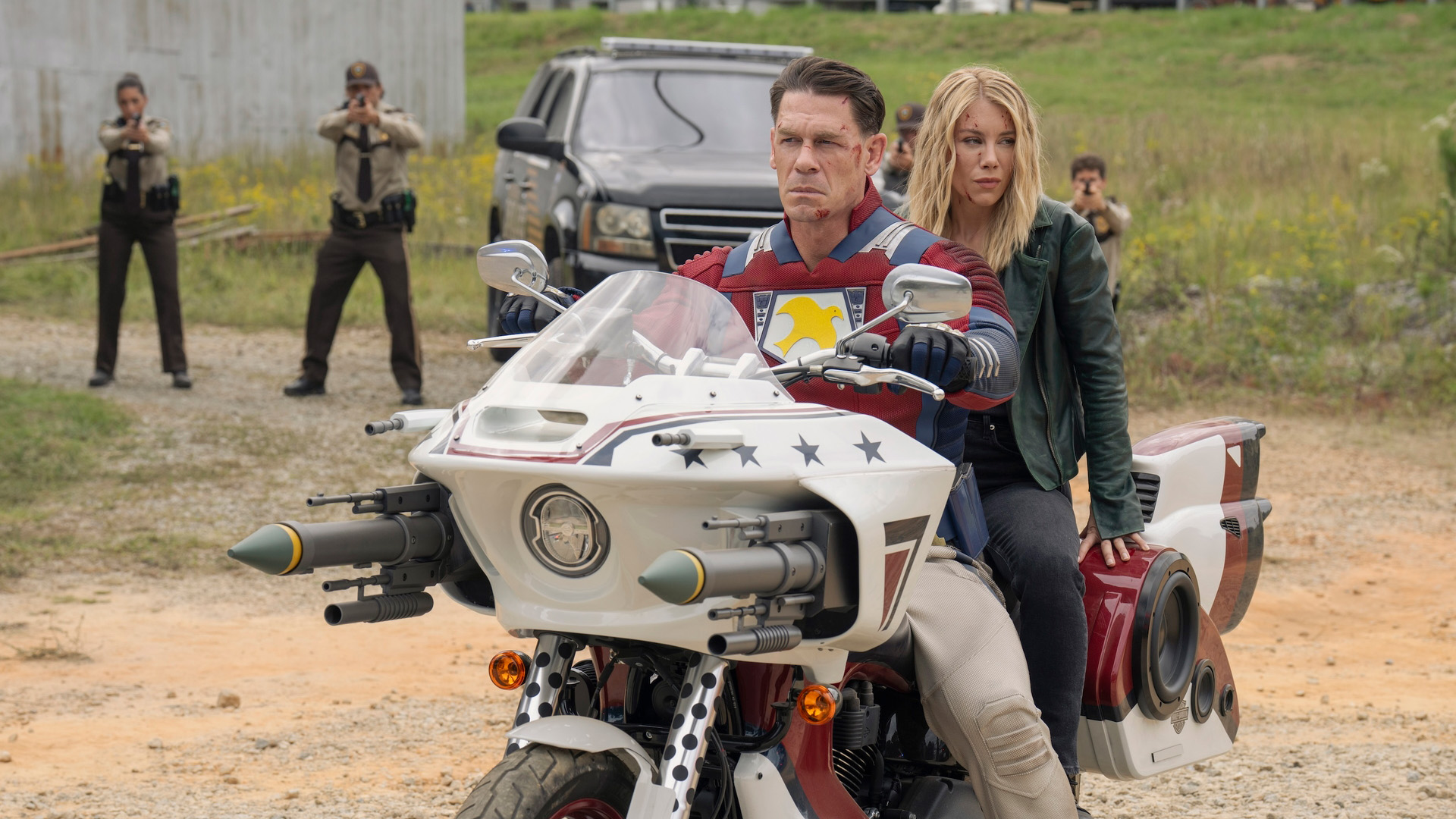 John Cena as Peacemaker and Jennifer Holland as Harcourt, surrounded by the police, in Peacemaker season 2.