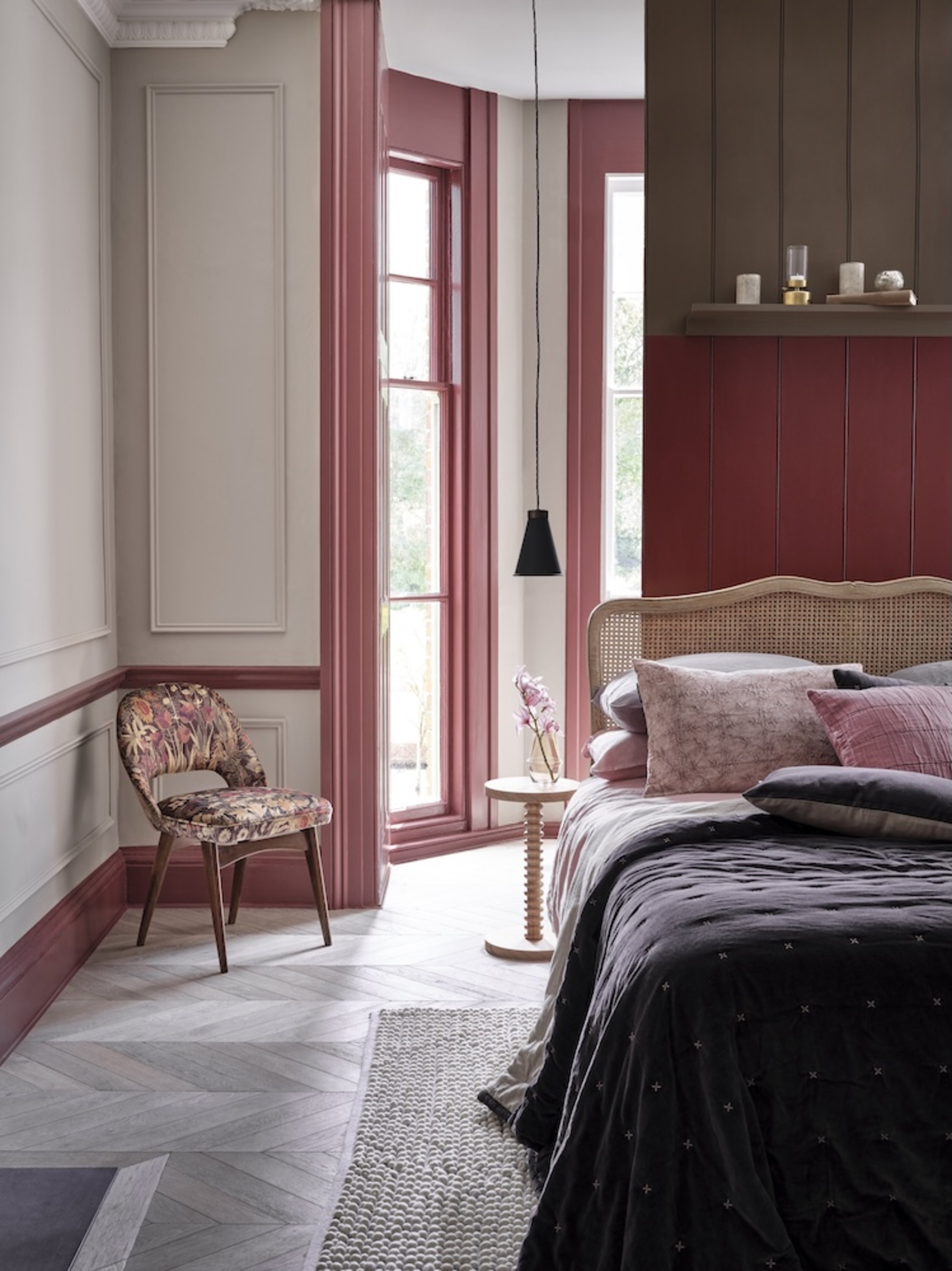 a bedroom with pink wall trims and molding details, a floral chair, a gray rug on top of parquetry flooring, and a floating bed against a paneled wall with a rattan headboard, lots of pillows, a dark bedcover, and a pink bobbin side table with a pendant light hanging overhead