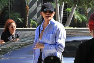 Chrissy Teigen wears black heeled ankle boots, black leggings, a striped button-down shirt, a black Gucci crossbody bag, a black hat, and a white sports bra.