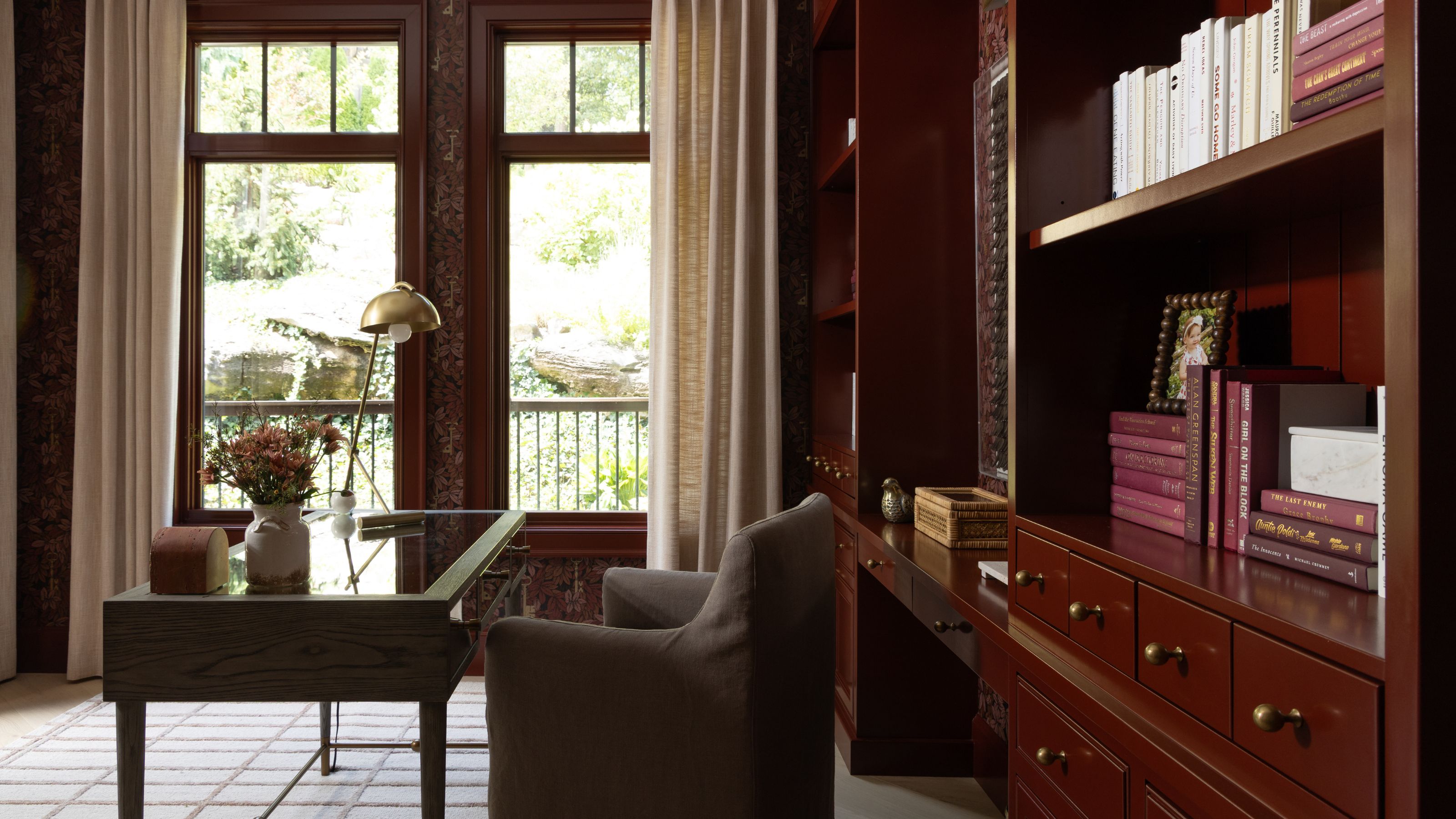 a dark red painted and wallpapered home office with custom cabinetry and tall windows overlooking the woods