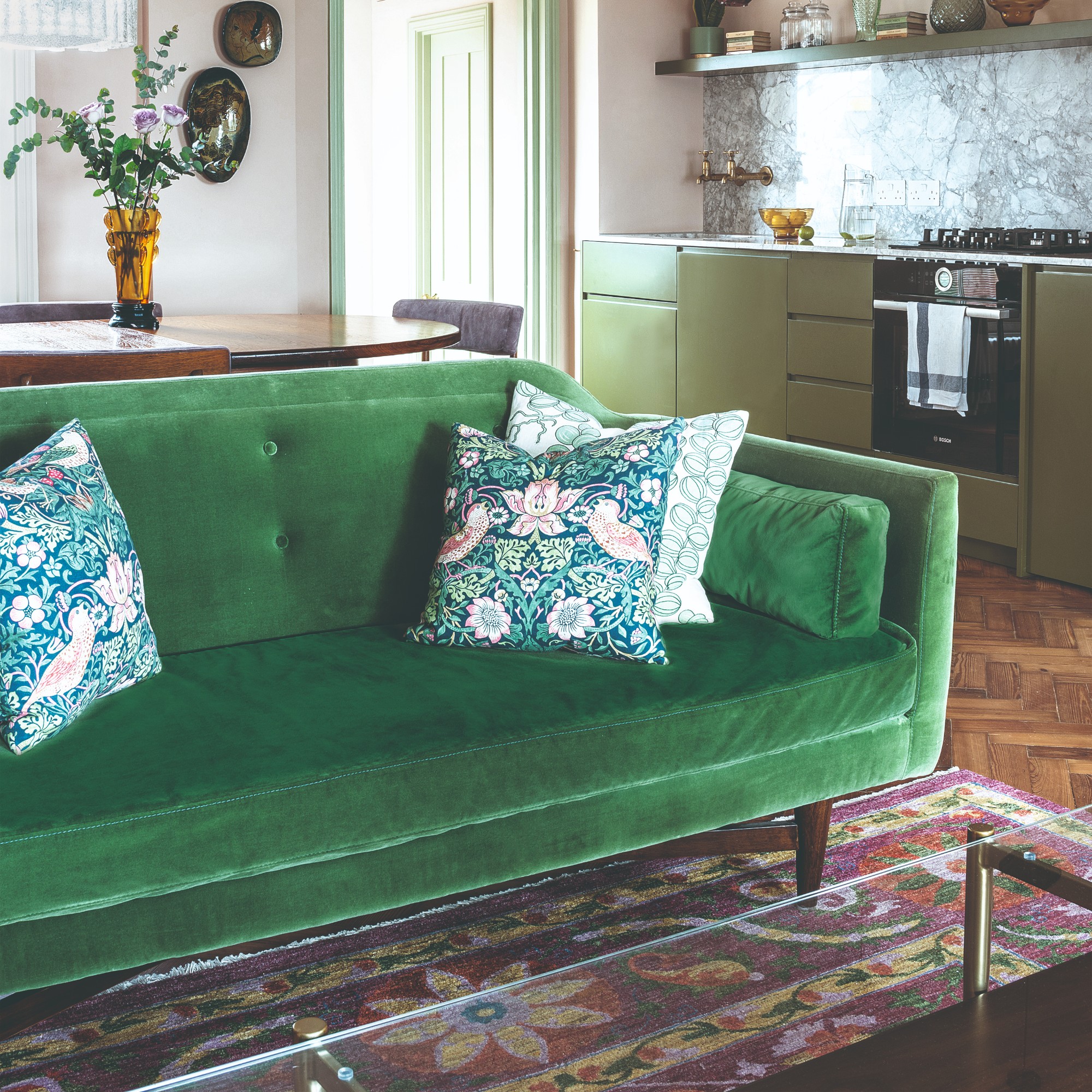 An open-plan living room with a midcentury-style green velvet sofa with floral-print scatter cushions and a floral rug underneath