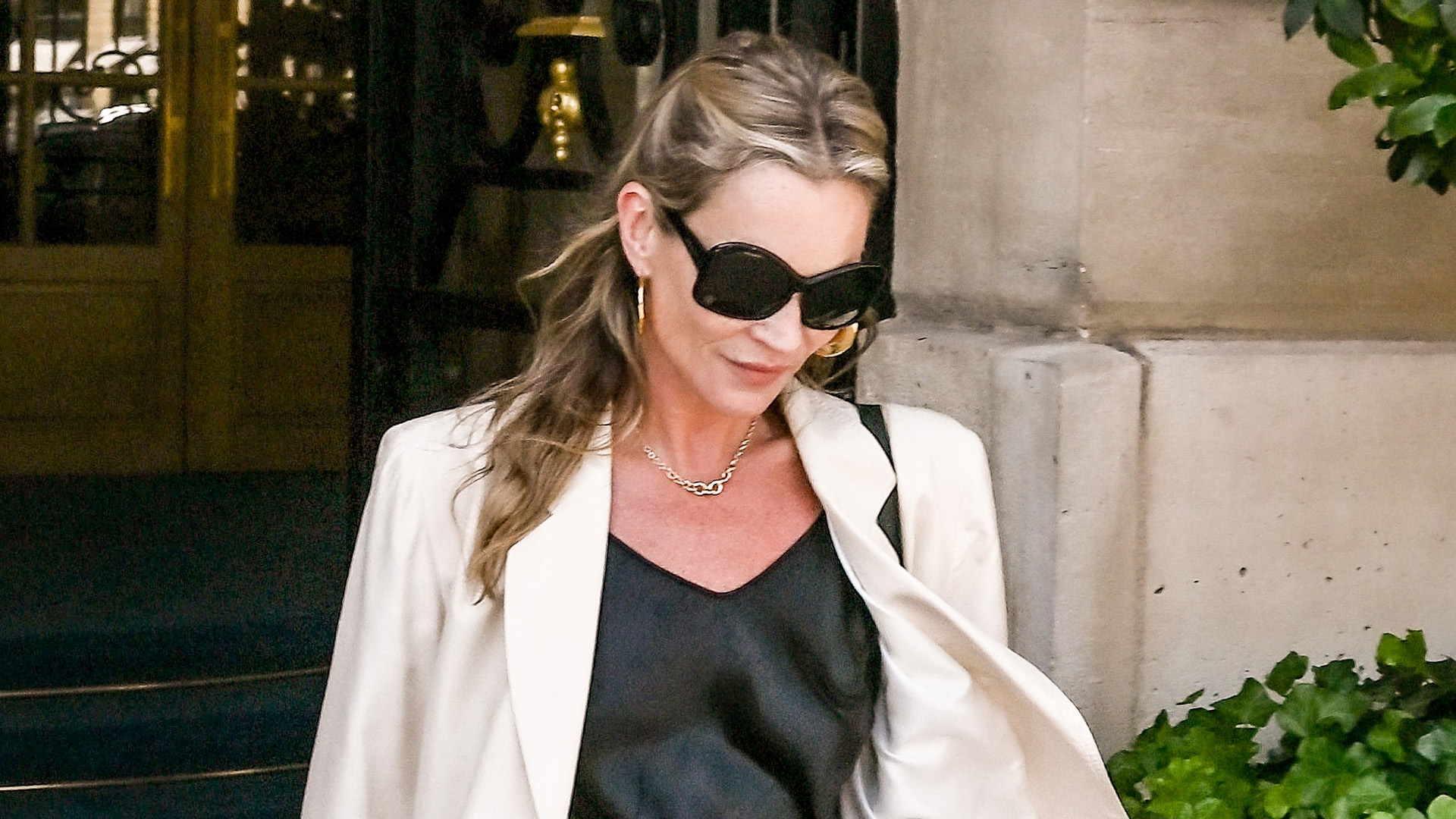 Kate Moss wearing a cream blazer, black slip dress, and ballet flats while exiting a hotel in Paris.