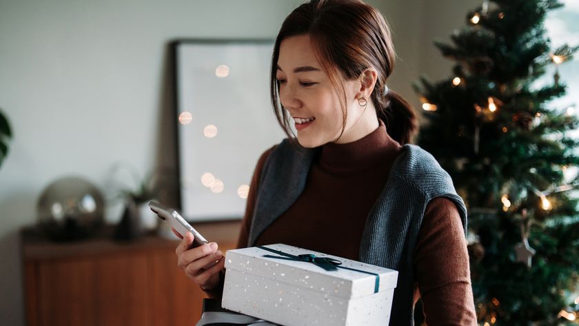 A smiling woman using smartphone while holding wrapped Christmas presents, standing against decorated Christmas tree.