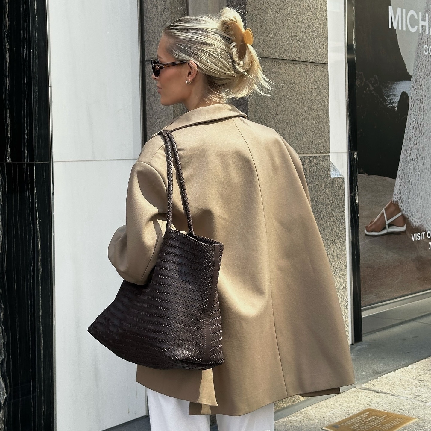 style influencer Fredrika Galgano poses in NYC wearing sunglasses, a camel claw clip, tan jacket, and brown woven leather tote bag