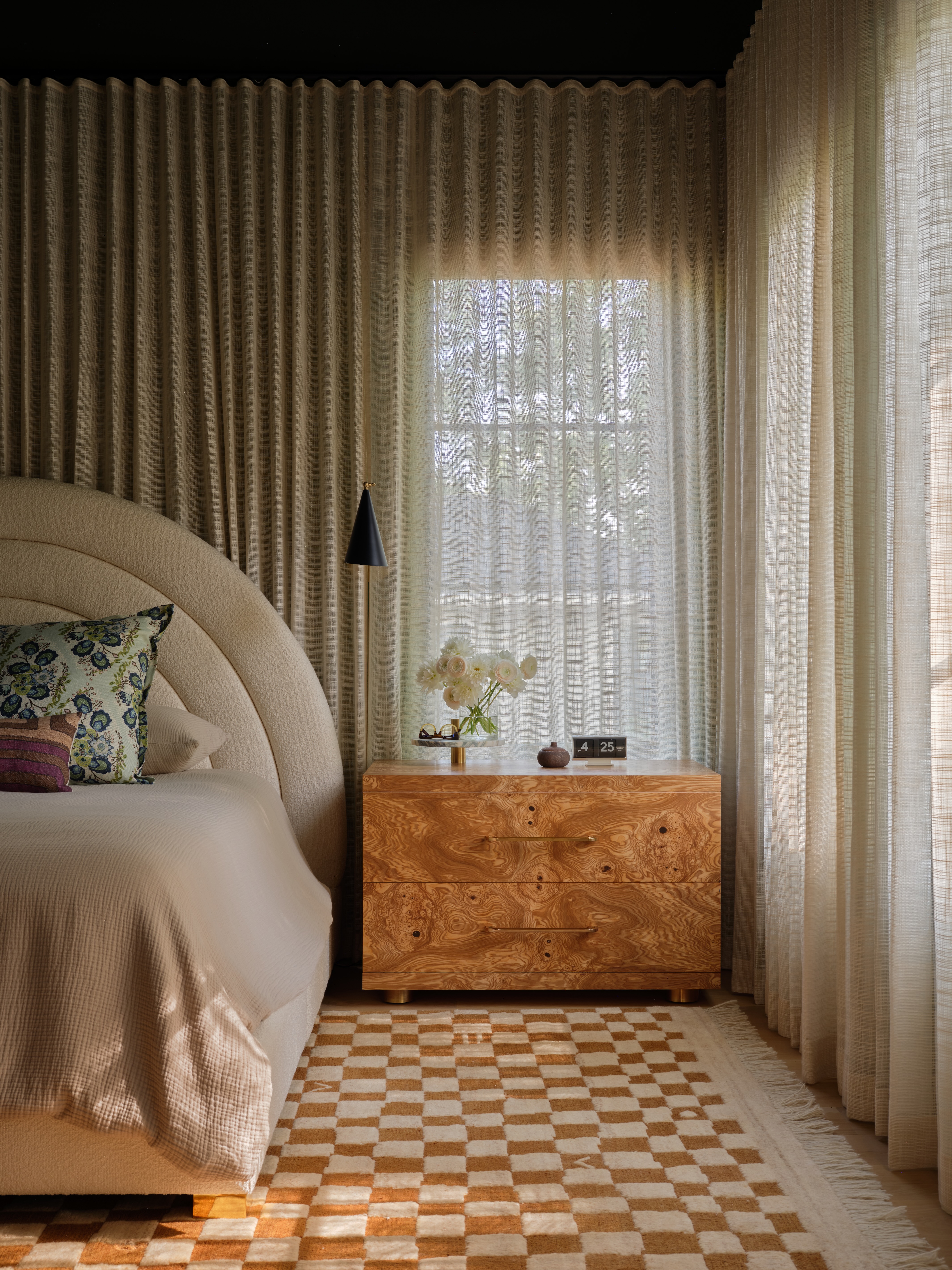 Sheer curtains wrap wall-to-wall in a bedroom with a rounded headboard atop a checkerboard rug.