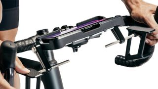 A close up of the new Kickr Steer, which is mounted to the front of a bike's handebars. It uses a black tray to hold a phone, and on either side, medal rods hold paddles out near the rider's thumbs