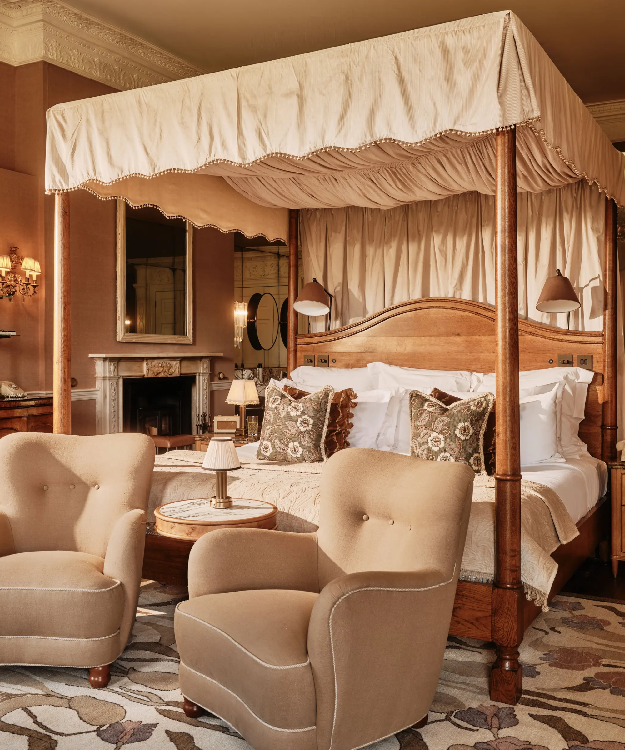 Richly layered bedroom featuring a a pair of piped-edge tufted armchairs, a wooden four-poster bed covered with a beige canopy, and an abundance of throw pillow in various sized, patterns, and colors.