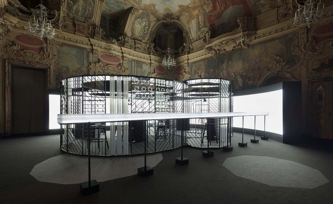 A clock-making workshop set up in the Milan’s Palazzo Visconti. The workshop is surrounded by harsh reflector lights, while the rest of the space is dark.