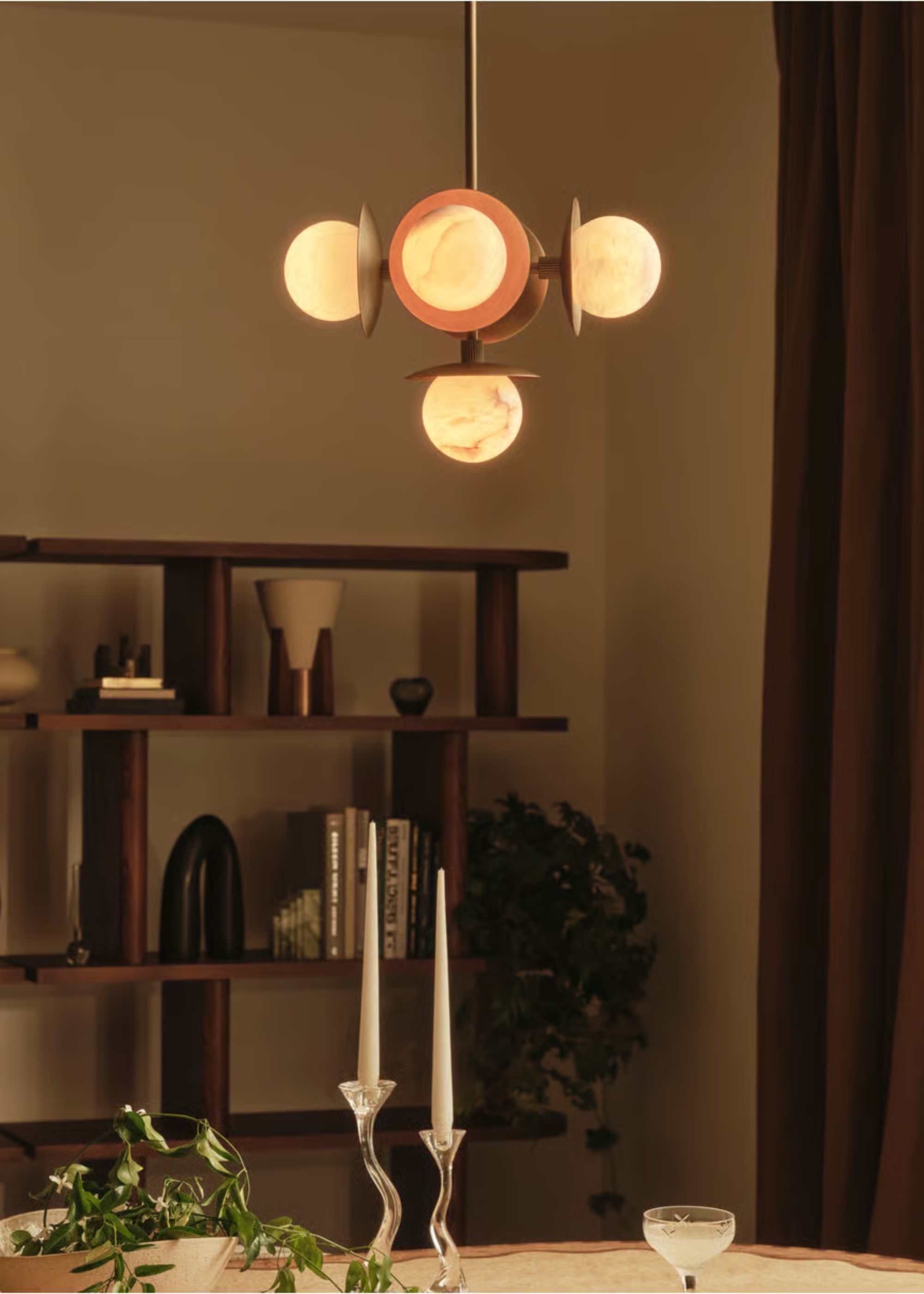 A pendant light hanging above a dining table that has two candles on top of it with a plant and glassware. There is also a view of a book shelf with decor pieces and books on top.