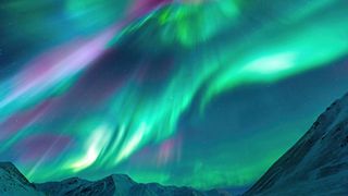 ribbons and curtains of light in the sky colored green, purple and pink. There are snow-capped mountains below. 